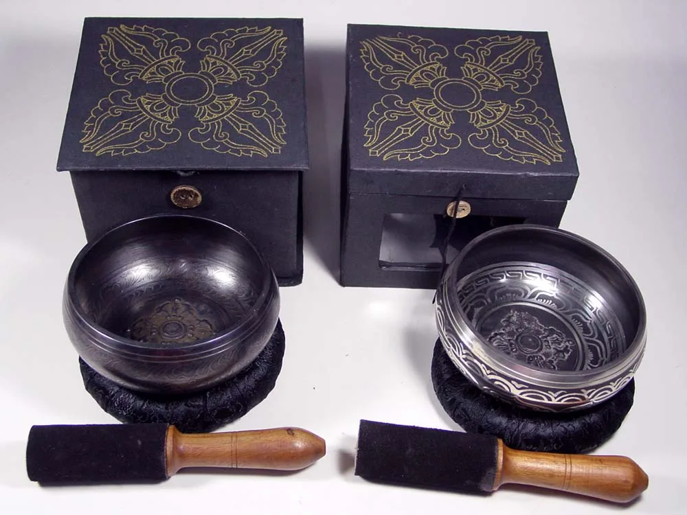 Two black singing bowls on black cushions with wooden mallets, in front of black gift boxes printed with a gold double dorje
